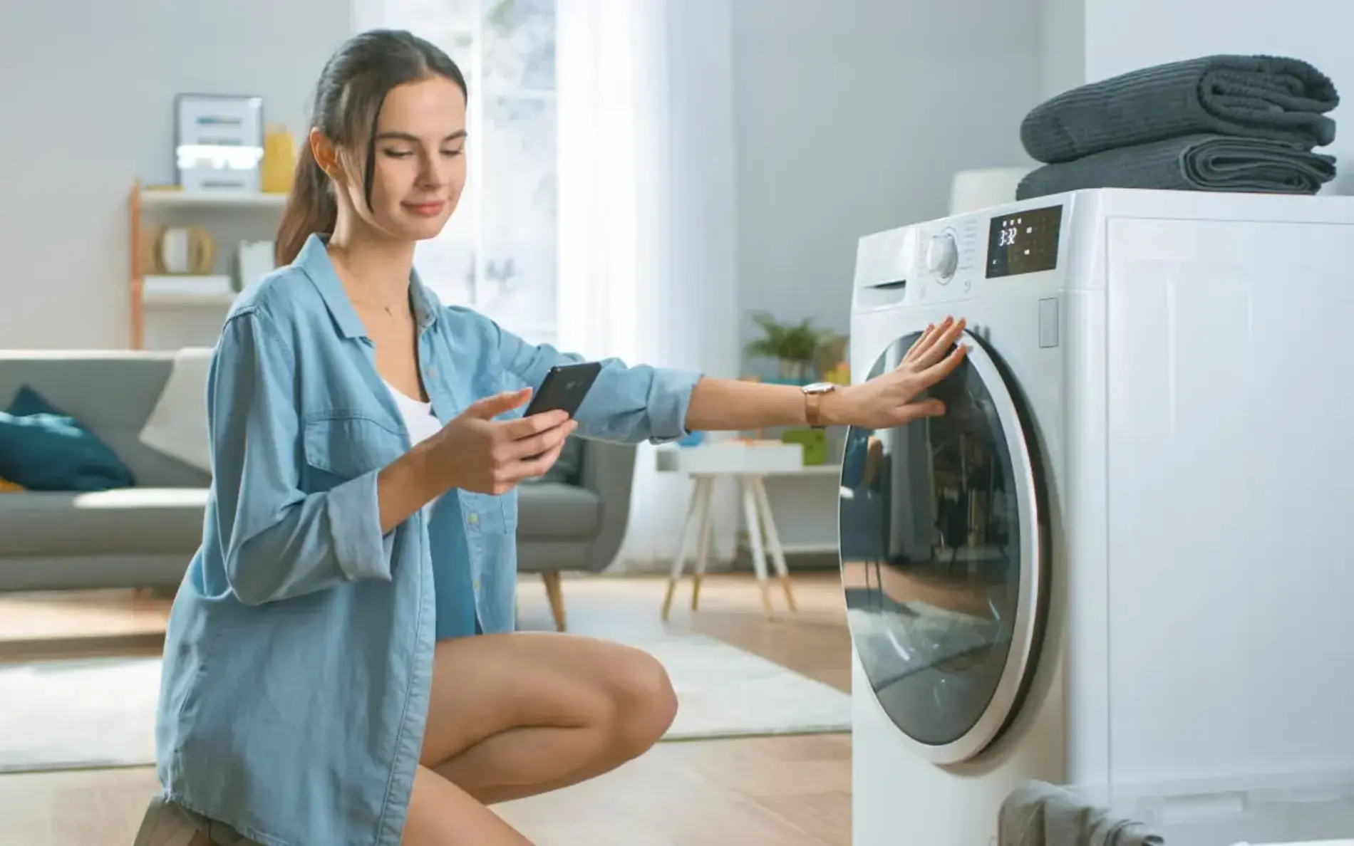 Eine Frau hockt neben ihrer Waschmaschine und schaut gleichzeitig auf ihr Handy und macht die Tür zu