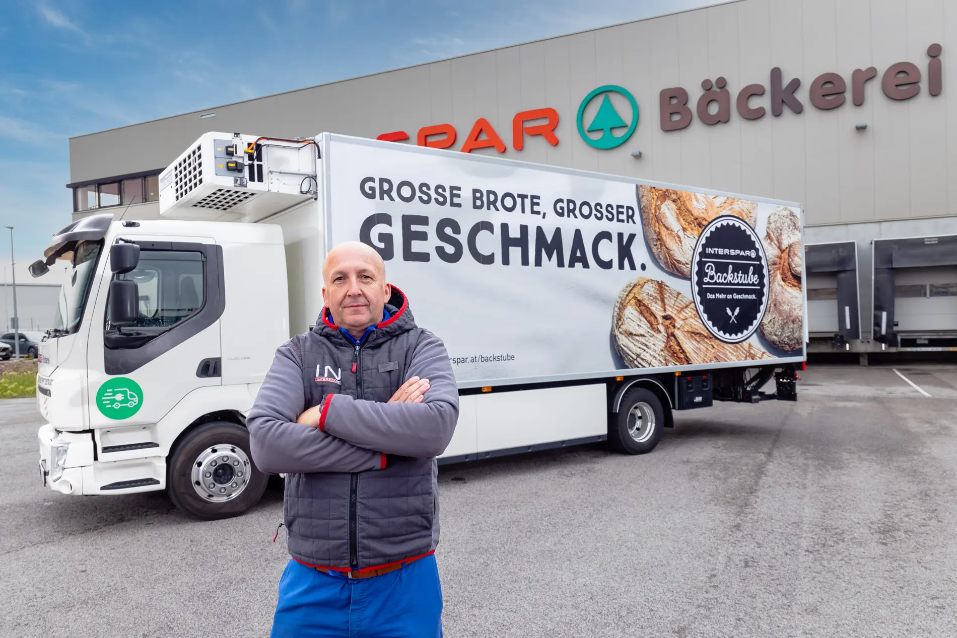 Mann steht mit verschränkten Armen vor einem E-Lkw von Spar. Dieser steht am Parkplatz vor der Spar Bäckerei Halle.