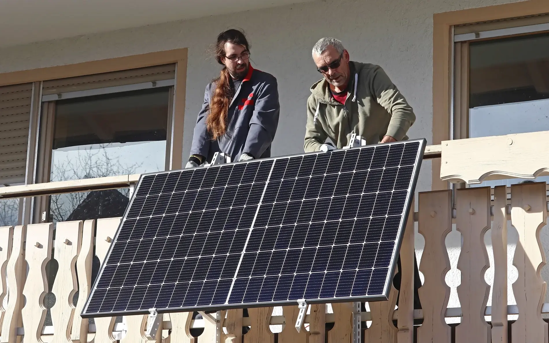 Zwei Männer installieren ein Balkonkraftwerk. Das Solarpanel ist in unsere Richtung ausgerichtet. Der Balkon ist aus Holz.