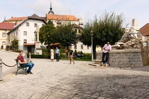 Ein öffentlicher Platz einer kleineren Gemeinde. Der Platz ist gepflastert. Links sitzt eine Person auf der Bank, rechts lehnt eine Person an der Wand und weiter hinten in der Mitte unterhalten sich zwei Personen.