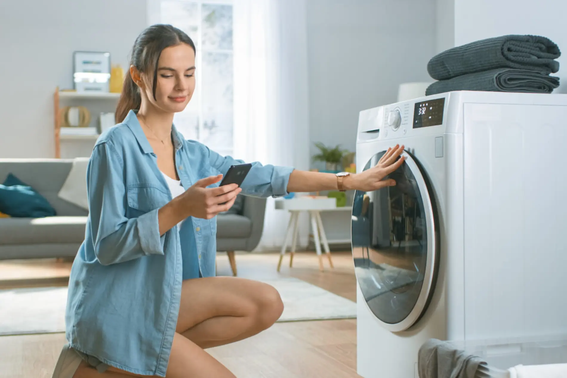 Eine Frau kniet vor der Waschmaschine hat ein Handy in der hand und macht mit der anderen Hand die Waschmaschinentür zu.