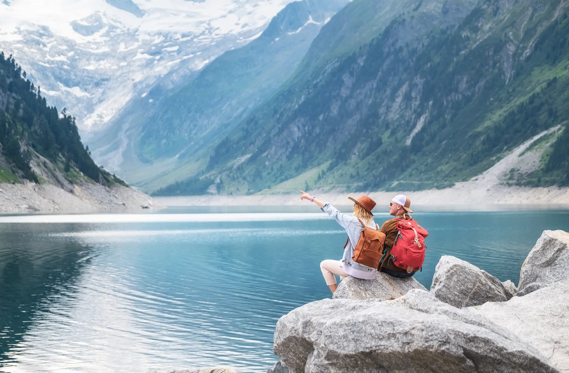 Zwei junge Menschen mit Rucksack sitzen am Fels. Vor Ihnen ein See umschlossen von Bergen.