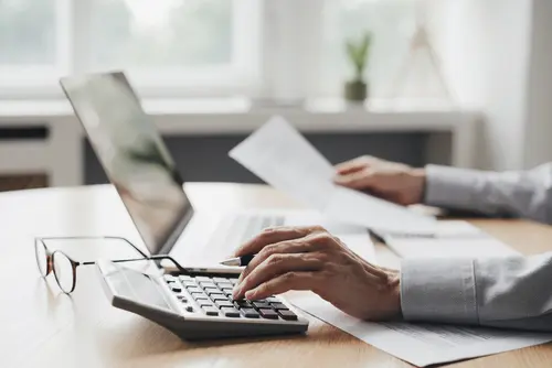 Eine Hand bedient einen Taschenrechner, gleichzeitig wird in der anderen Hand ein Blatt gehalten. Die Person sitzt vor einem Laptop und die Brille liegt am Schreibtisch.