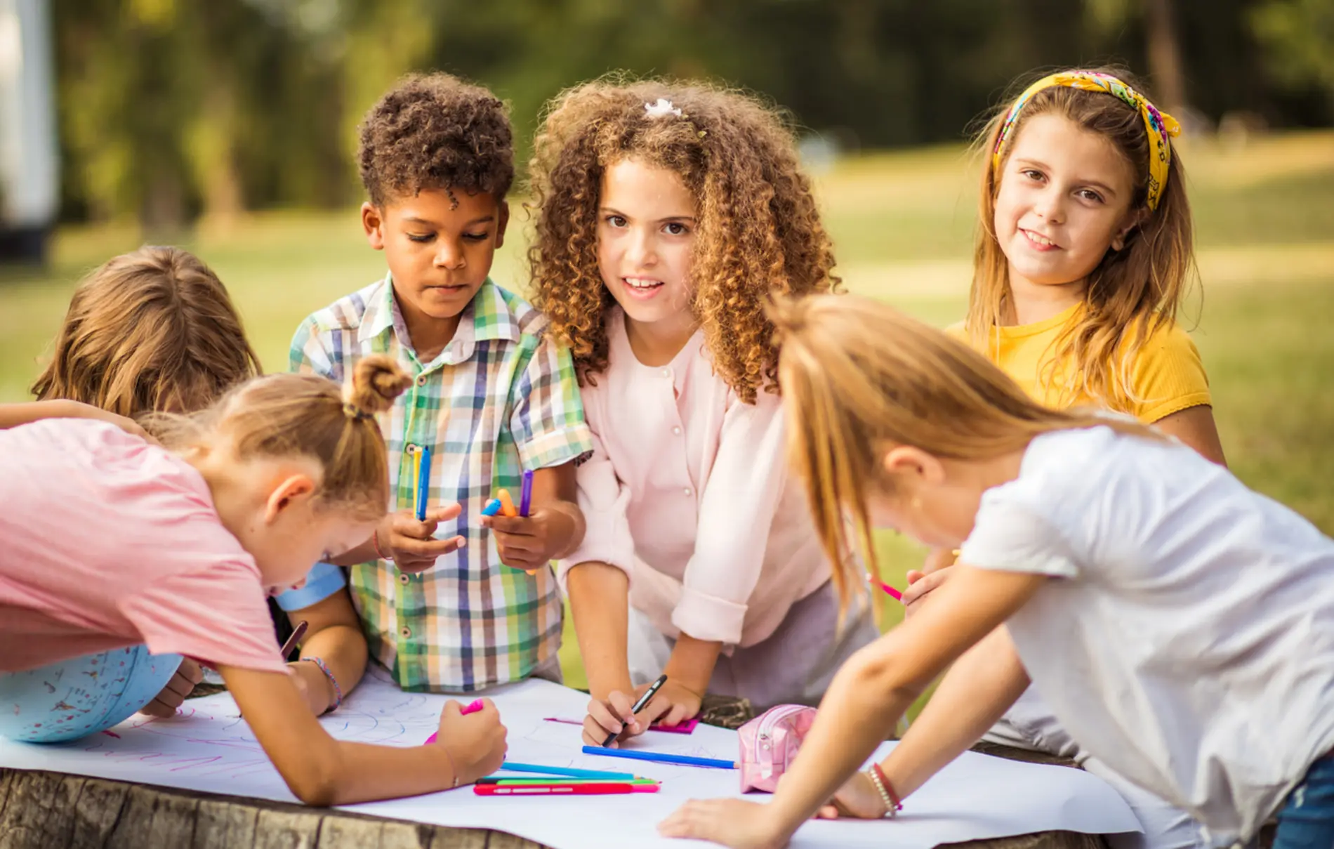 Kinder sitzen gemeinsam im Freien um einen Holztisch herum und zeichnen auf Papier