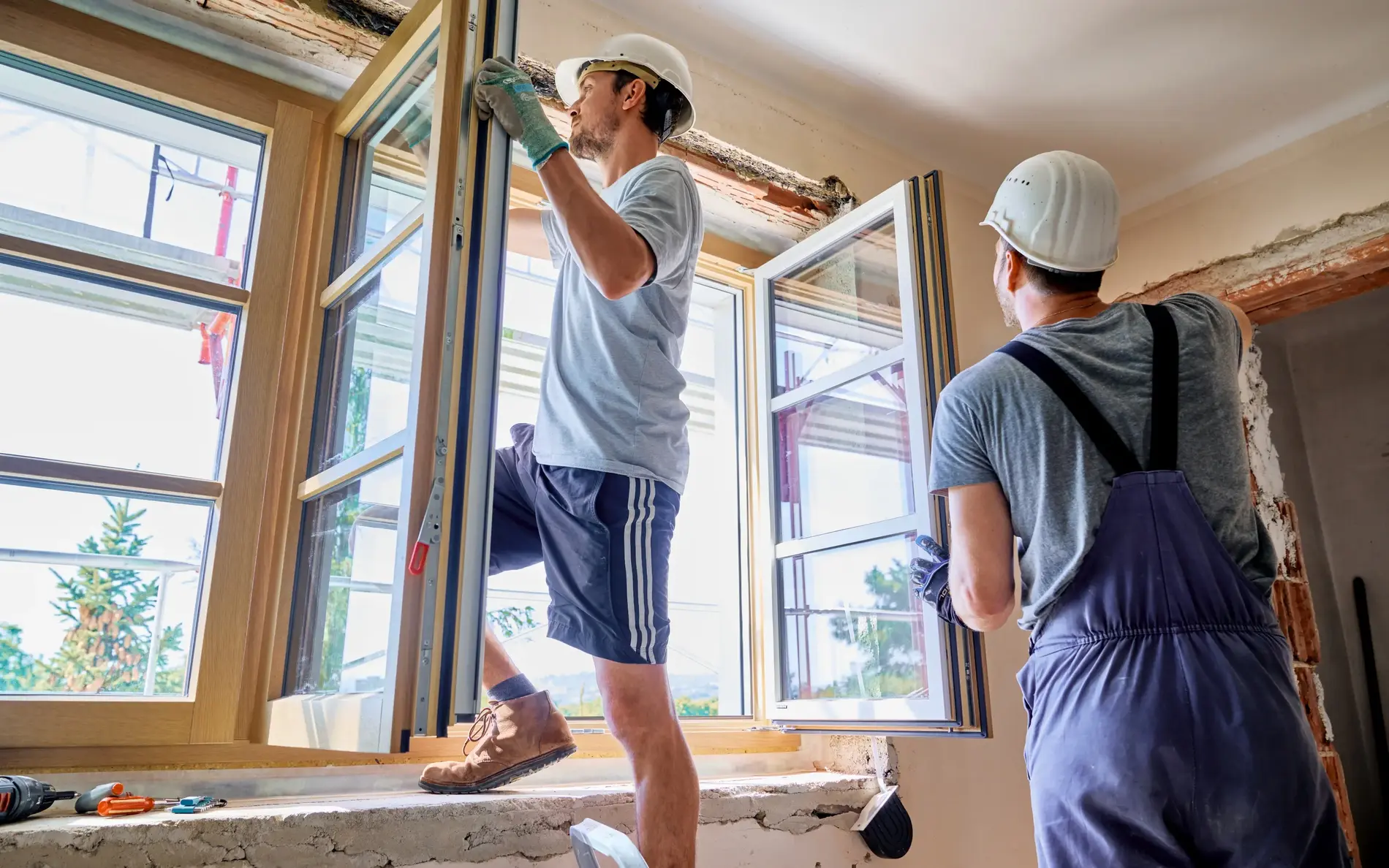 Zwei Männer bauen gerade Fenster ins Haus ein. Ein Mann steht am Fensterbrett und fixiert das Fenster, der andere schaut zu ihm hinauf.