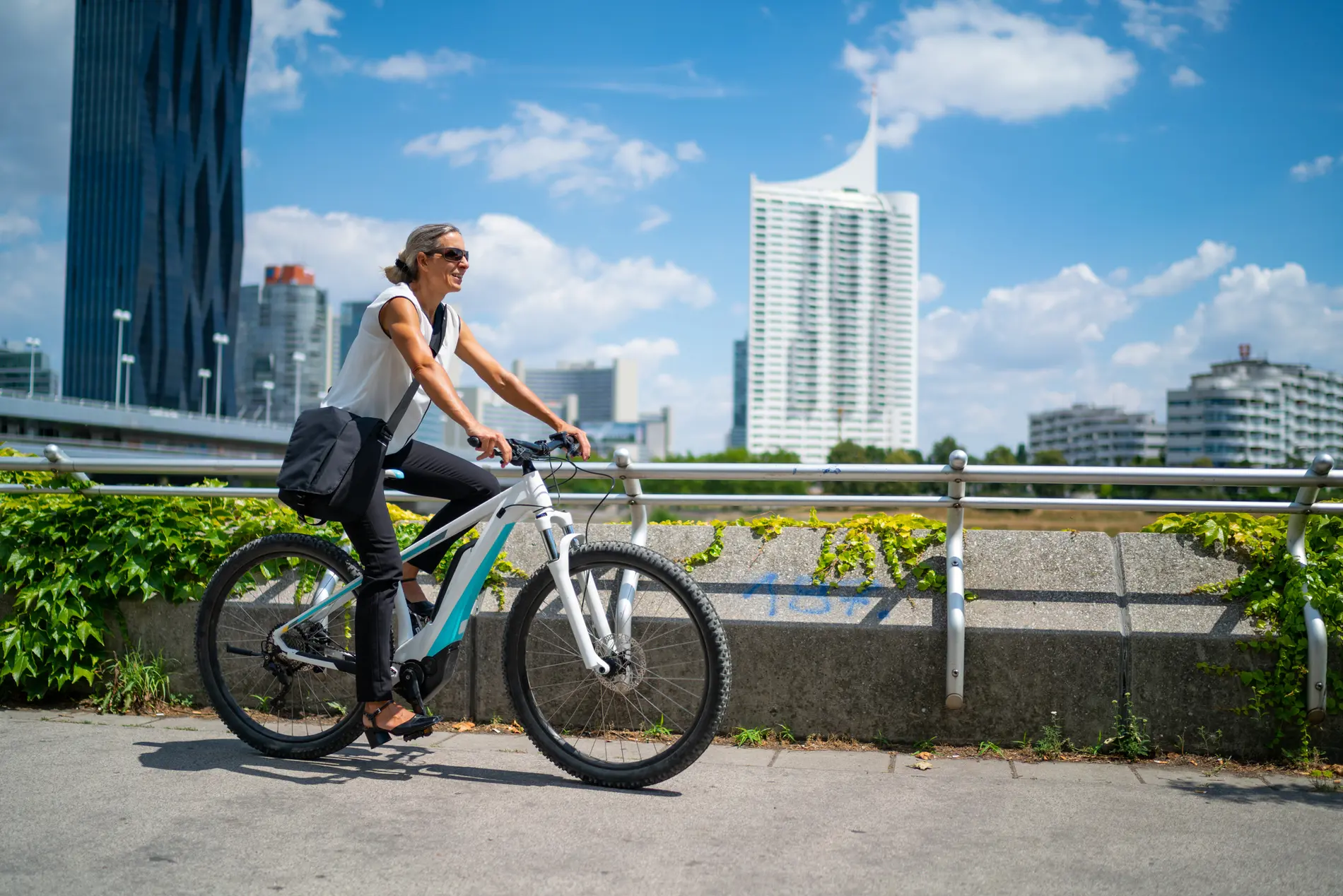 Eine blonde Frau mit Sonnenbrille fährt auf einem E-Bike, im Hintergund sind Hochhäuser und eine Brücke zu sehen.