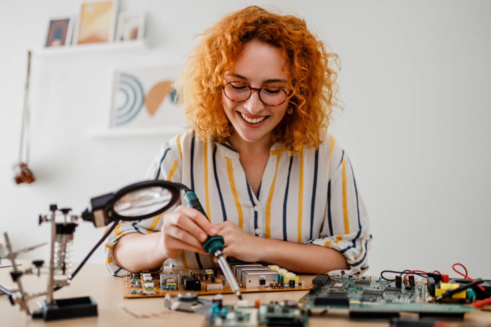 Zu sehen ist eine Frau mit rotem gelockten Haar, die auf einem Schreibtisch sitzt und Elektronikteile zusammenbaut.