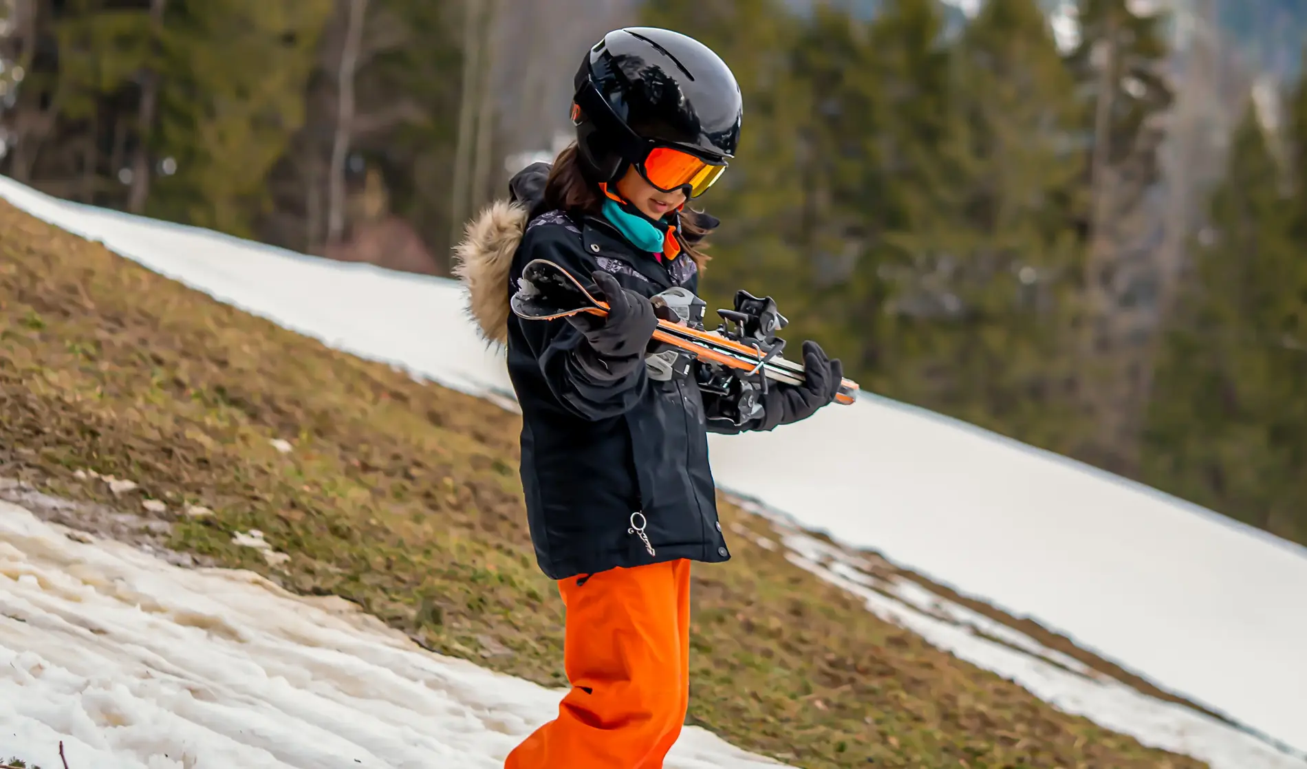 Ein Kind geht im Skianzug mit den Skiern in den Händen an einem kleinen weißen Schneeband hinunter. Im Hintergrund sieht man, dass die Skipiste in weiten Teilen grün ist.