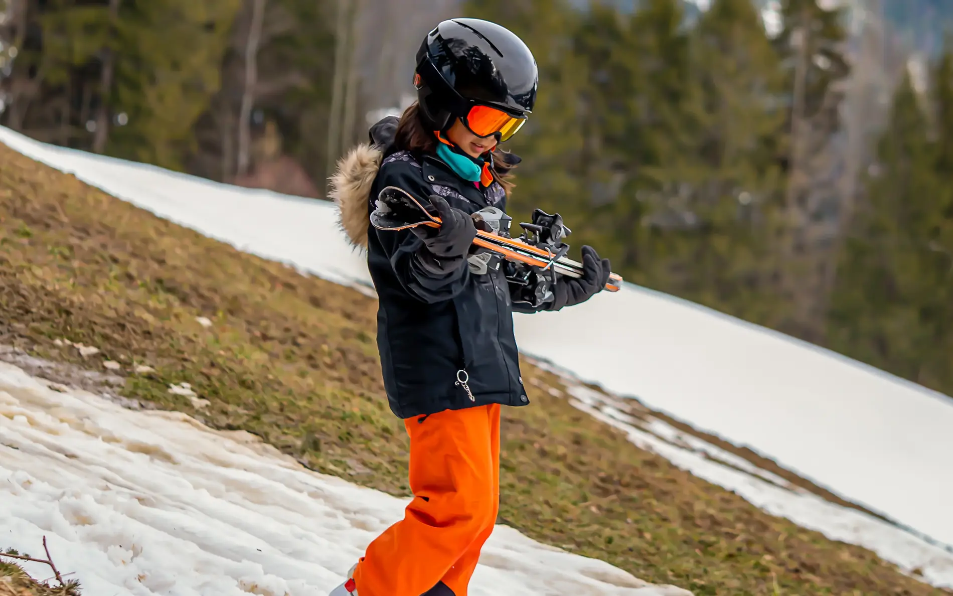 Ein Kind geht im Skianzug mit den Skiern in den Händen an einem kleinen weißen Schneeband hinunter. Im Hintergrund sieht man, dass die Skipiste in weiten Teilen grün ist.