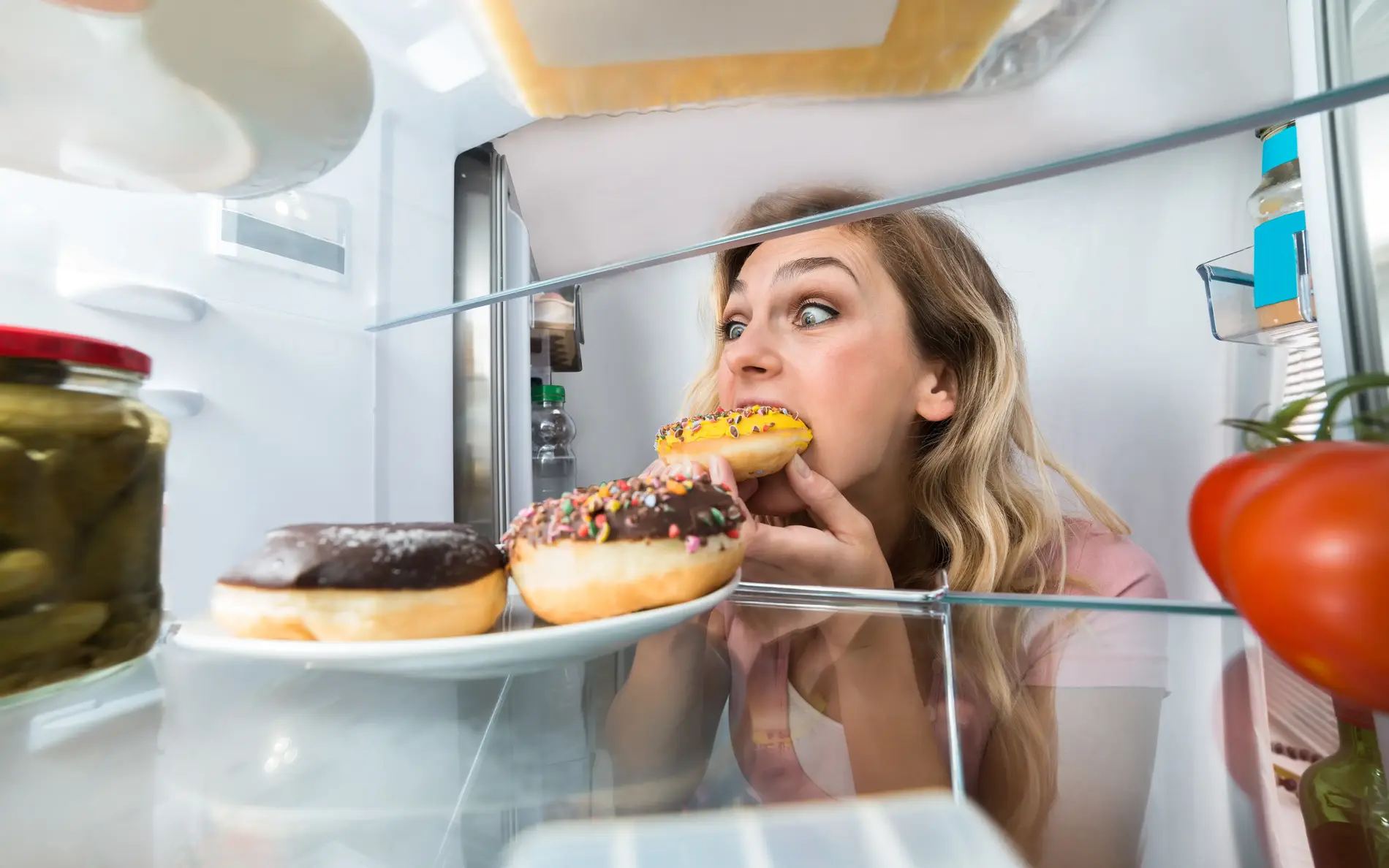 EIne blonde Frau isst noch an der Kühlschranktür einen Donut aus dem Kühlschrank.