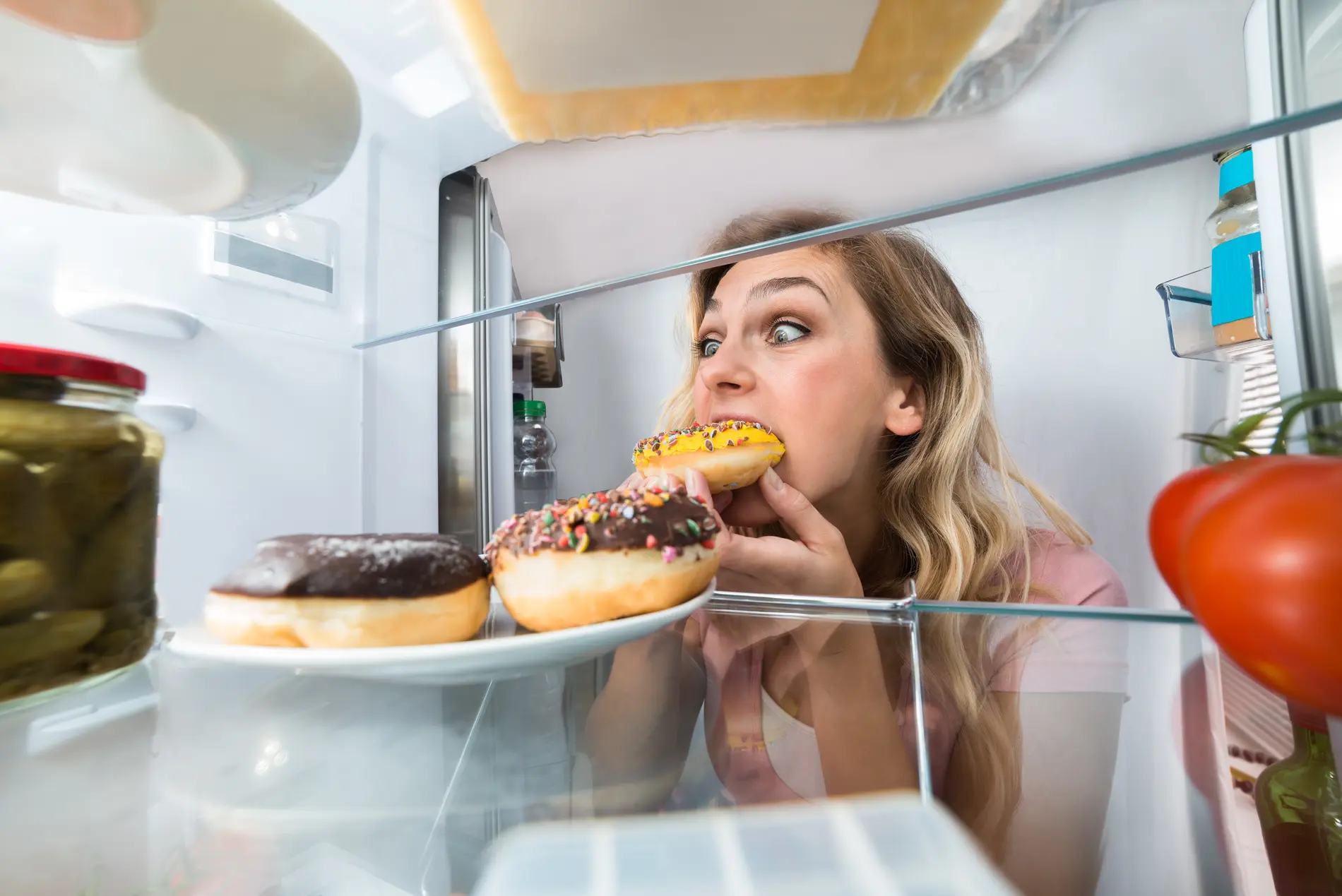 EIne blonde Frau isst noch an der Kühlschranktür einen Donut aus dem Kühlschrank.